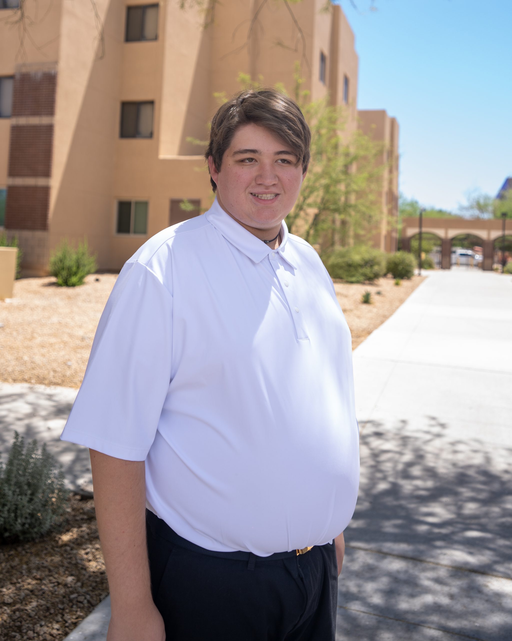 Close-up headshot of a teenage boy with brown hair in a white polo shirt, smiling slightly, outdoors on a sunny day.