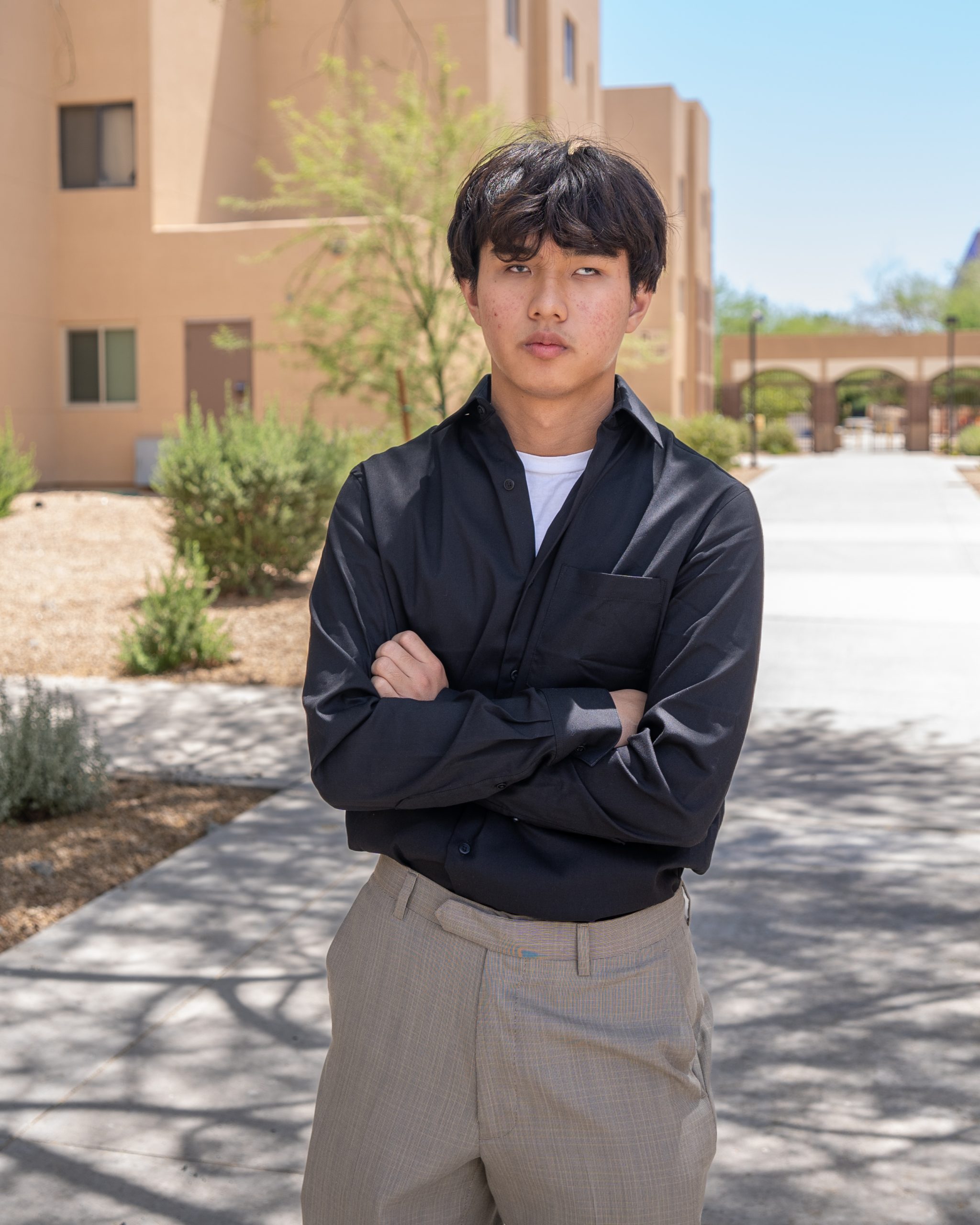 Full-body shot of a teenage boy with black hair, wearing a black shirt and beige trousers, standing confidently on a sunny campus walkway.
