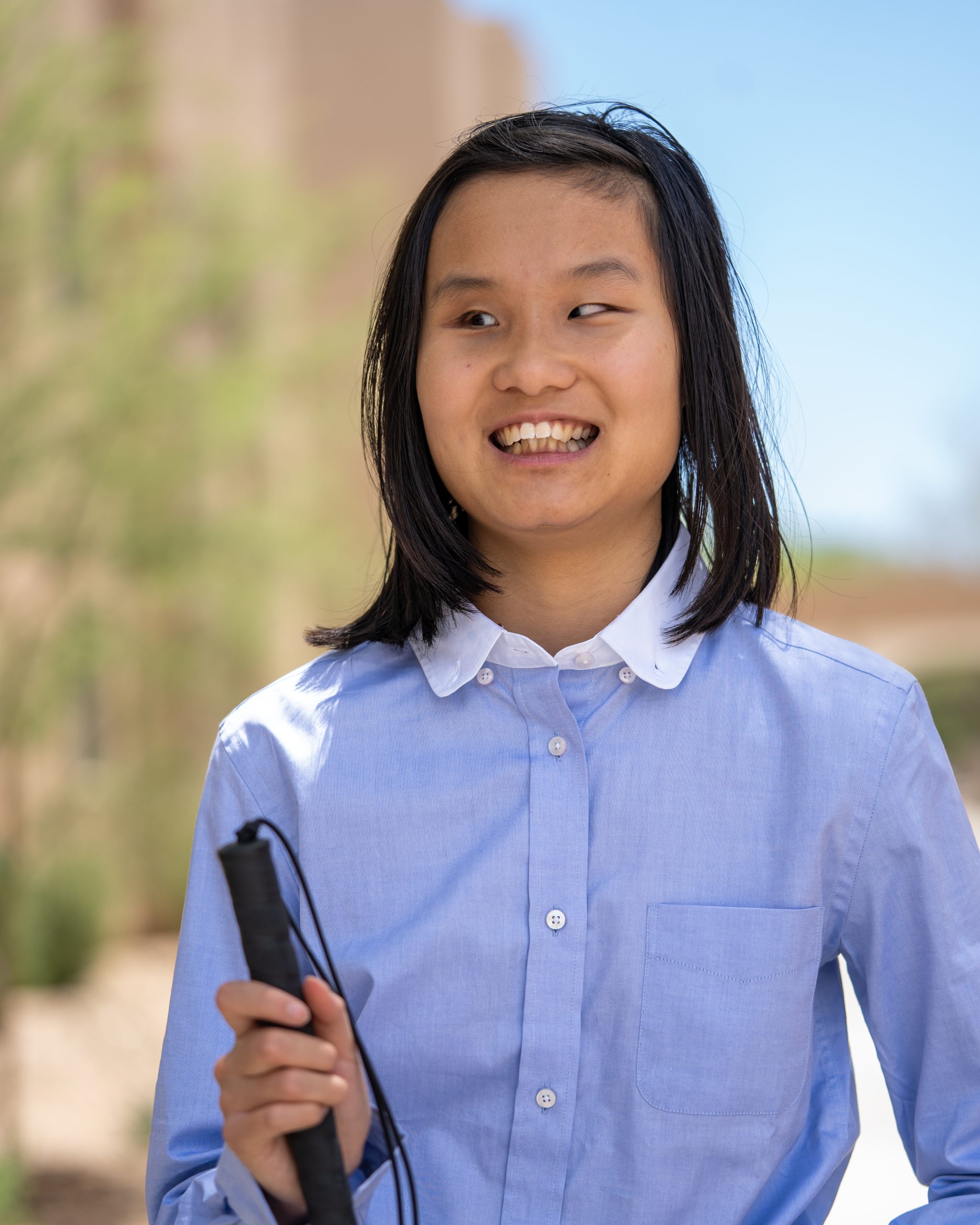 Girl smiling widely, posing with her new dress-for-success outfit, wearing a blue button-up shirt and black trousers, and holding her white cane.