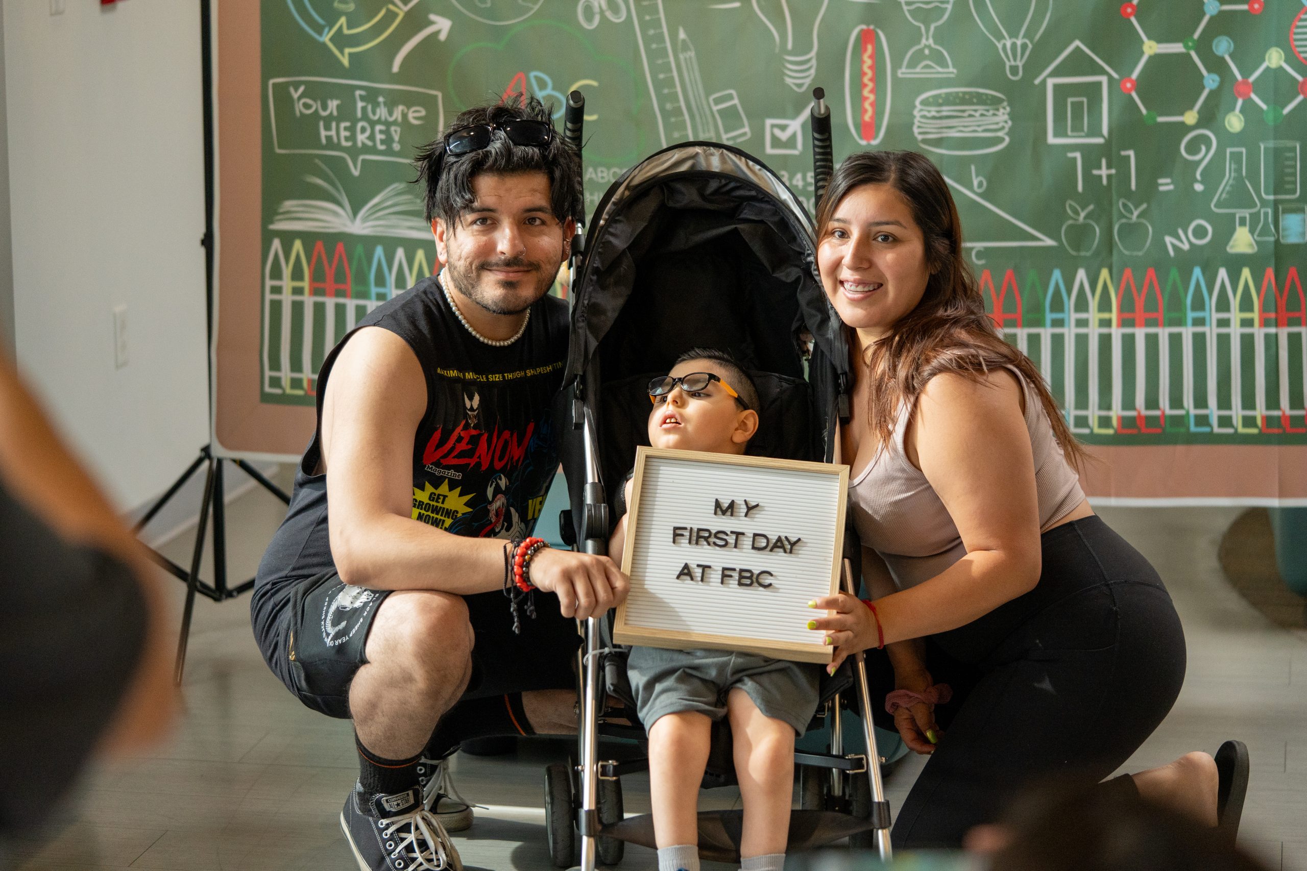 Parents and a young boy smile for the camera holding a board that reads My first day at FBC