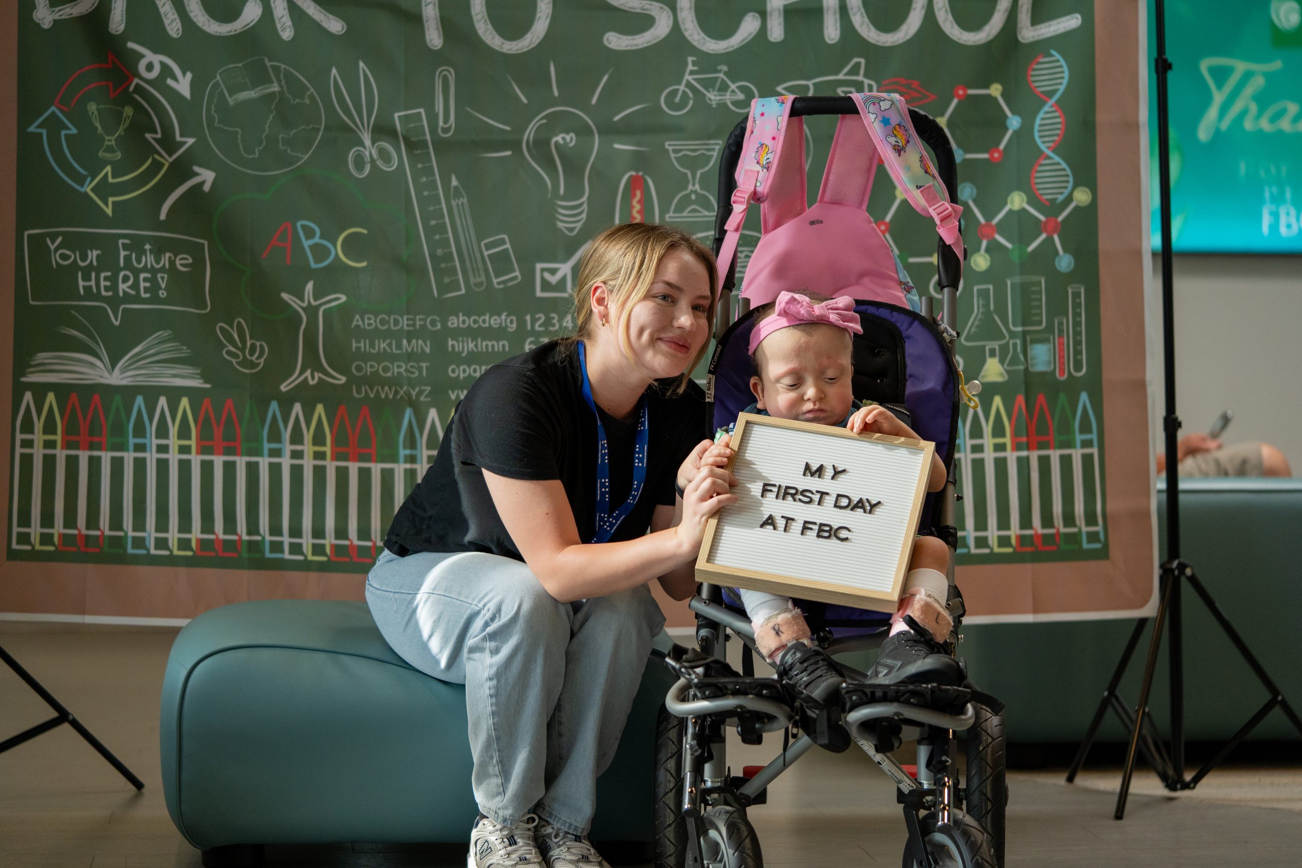 A teacher and a young child smile at the camera while holding a board that says 'My First Day at FBC.'