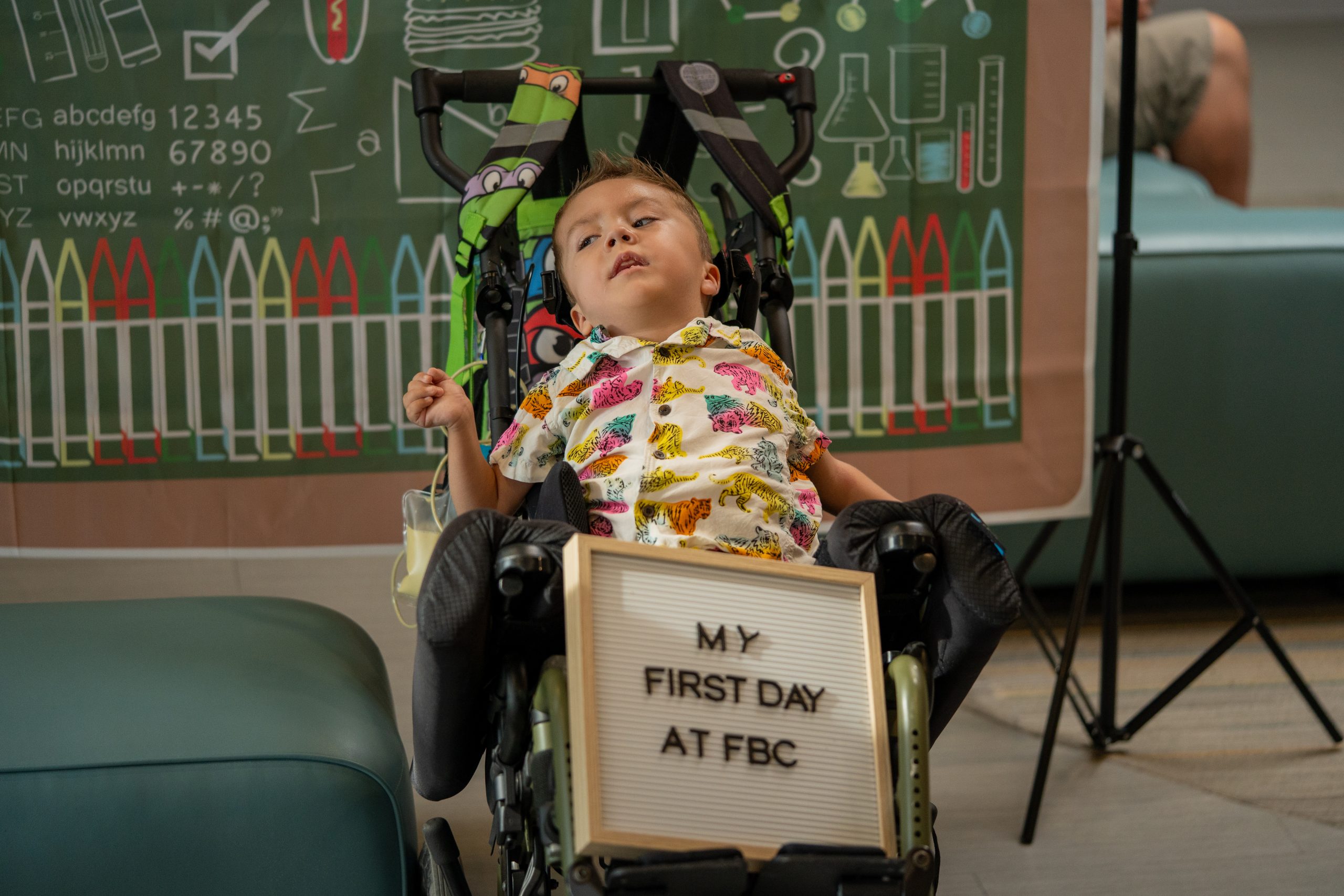 A young boy wearing a colourful shirt poses with a board that reads: My first day at fbc