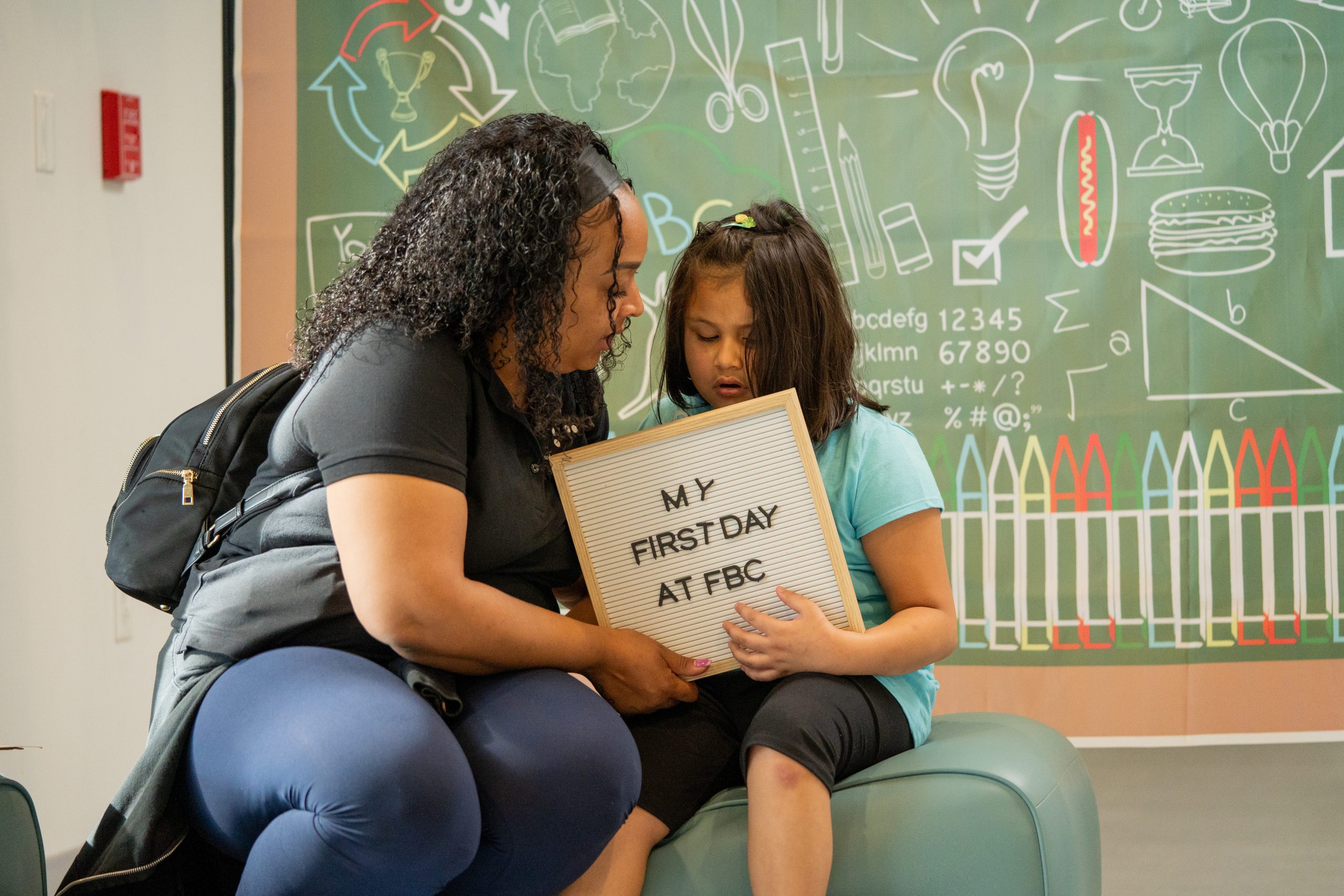 A teacher describes the contents of a board to a young girl. The board reads 'My First Day at FBC.
