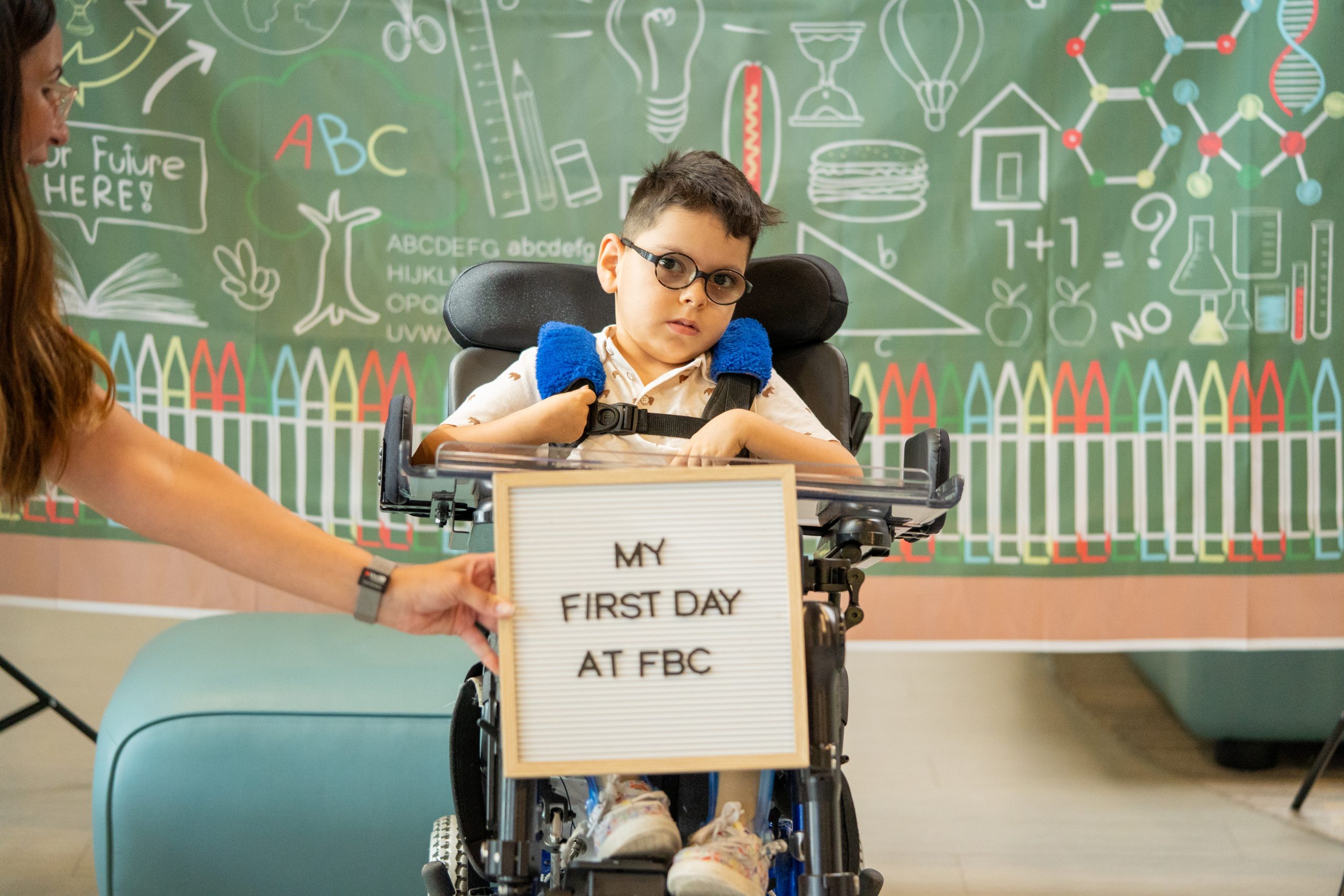 A young boy in a wheelchair wearing glasses smiles at the camera while someone holds a sign that says 'My First Day at FBC.'