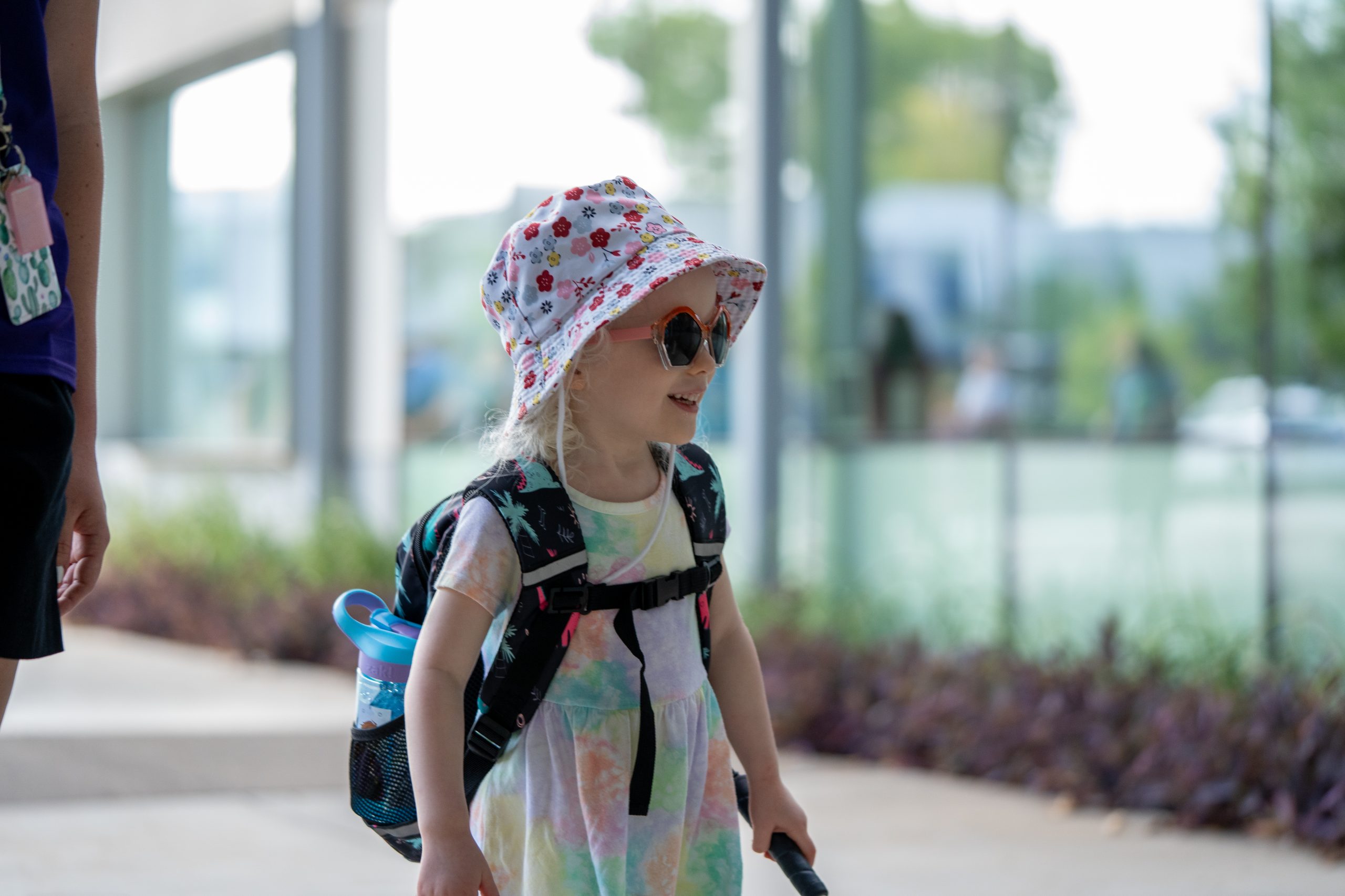 A young girl wearing a pink bucket hat and dark glasses holds her white cane and smiles