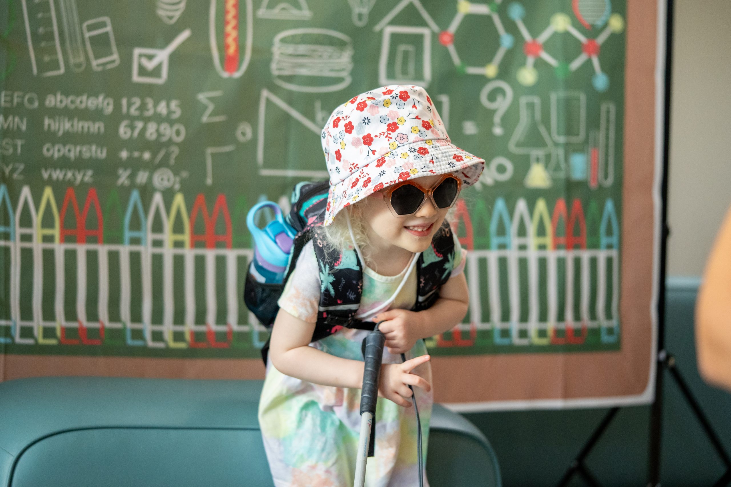 A young girl wearing a pink bucket hat and dark glasses holds her white cane and smiles