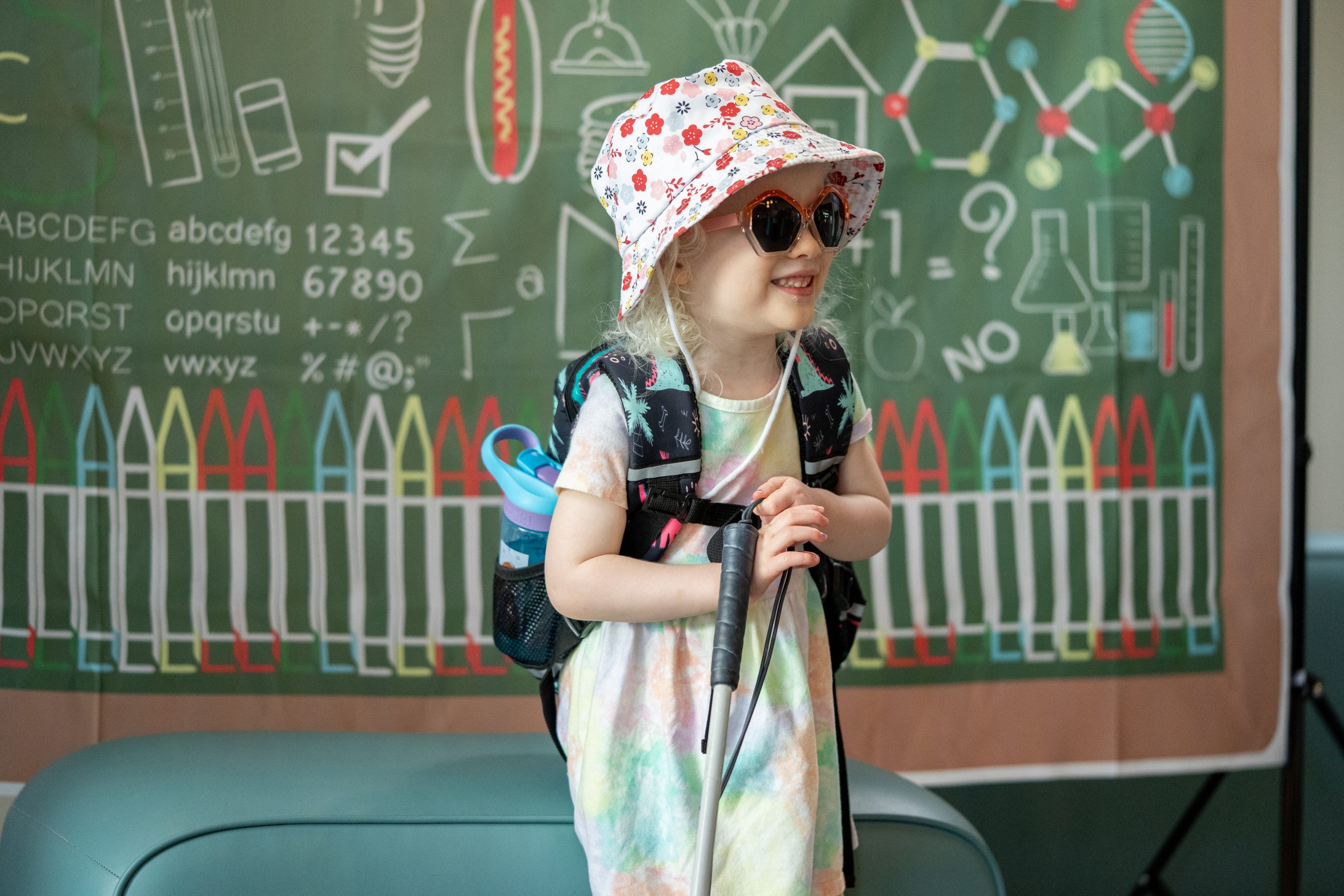 A young girl wearing a pink bucket hat and dark glasses holds her white cane and smiles