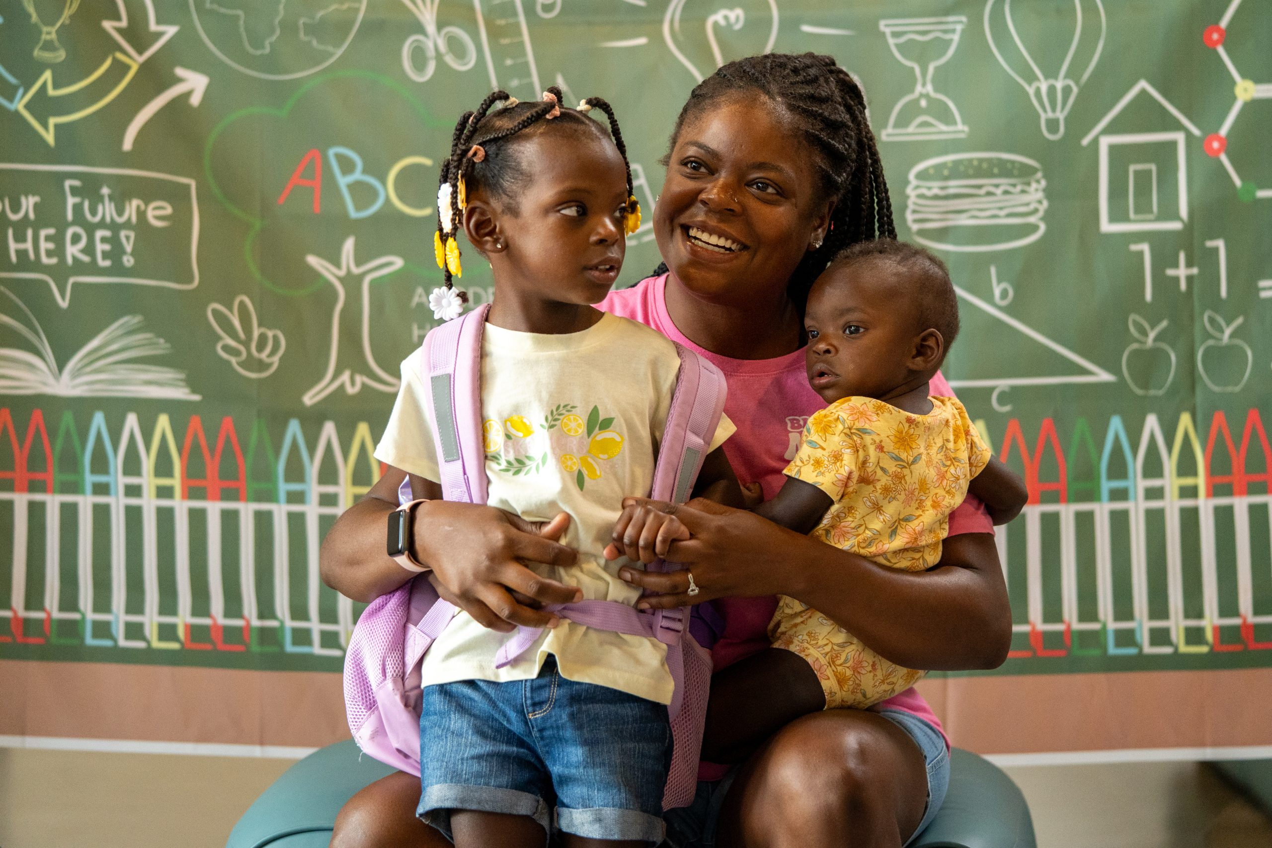 A mother smiles for the photo while hugging her young daughter and baby