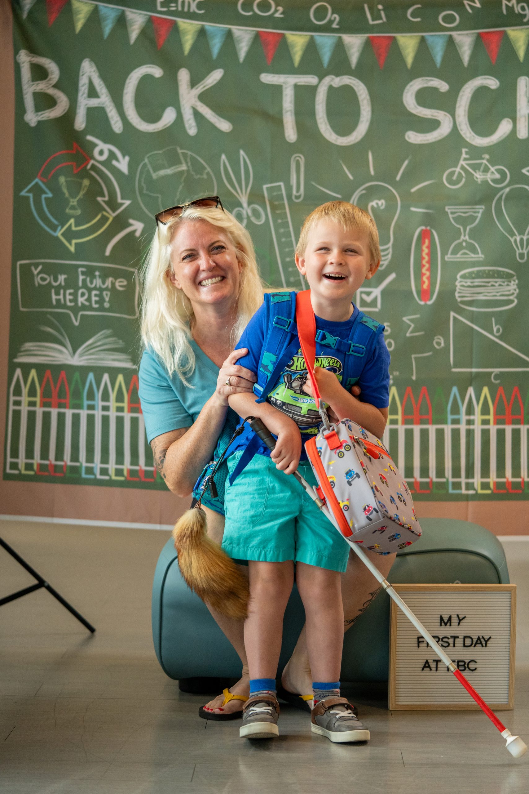 A young boy holding his backpack and a white cane smiles alongside his mother.