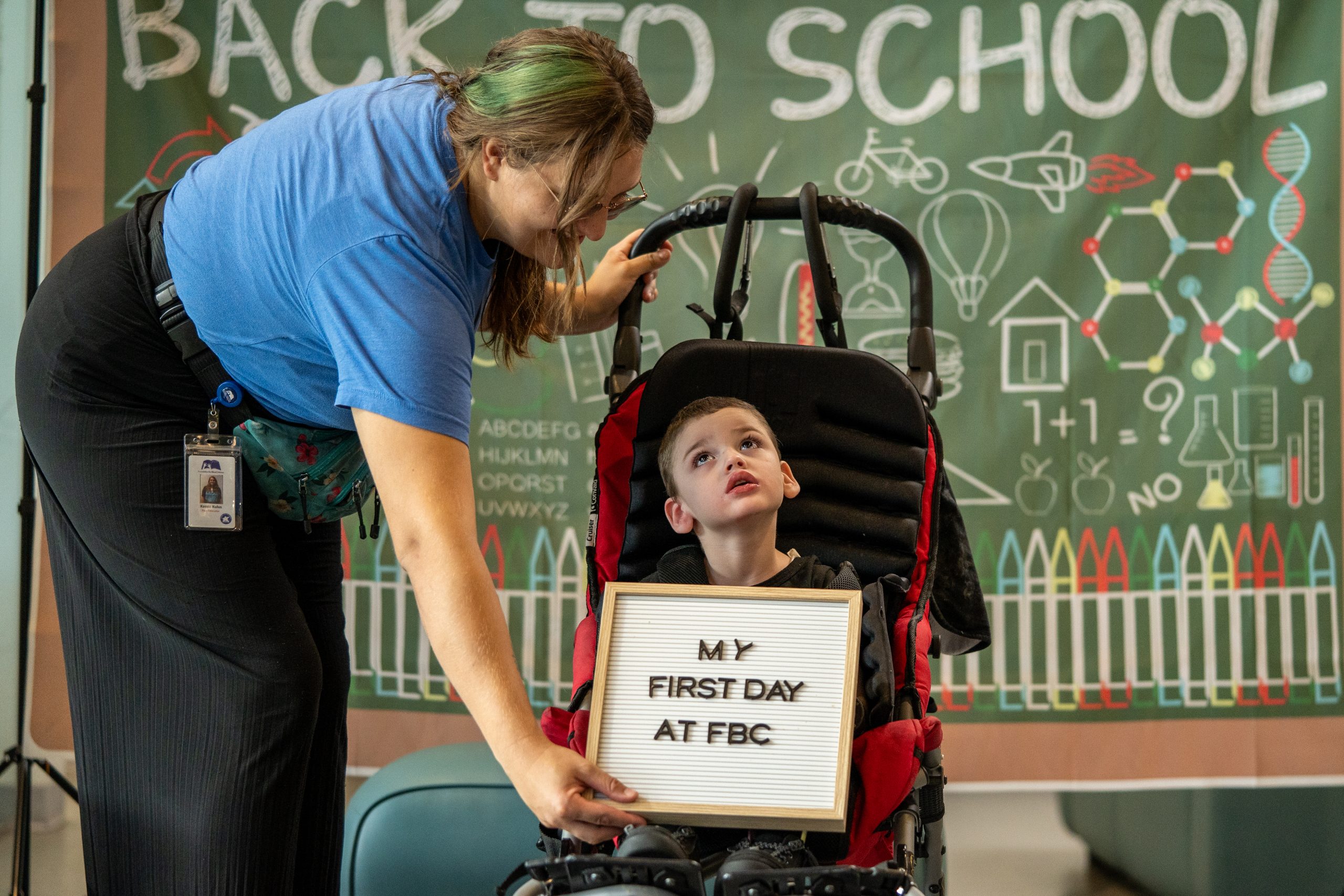 "A paraeducator smiles at the camera while holding a board that says 'My First Day at FBC' next to a little boy.