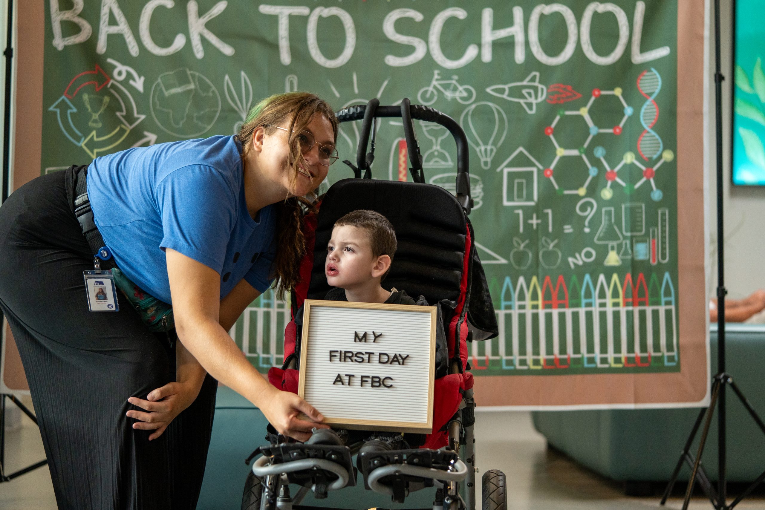 A paraeducator smiles at the camera while holding a board that says 'My First Day at FBC' next to a little boy.